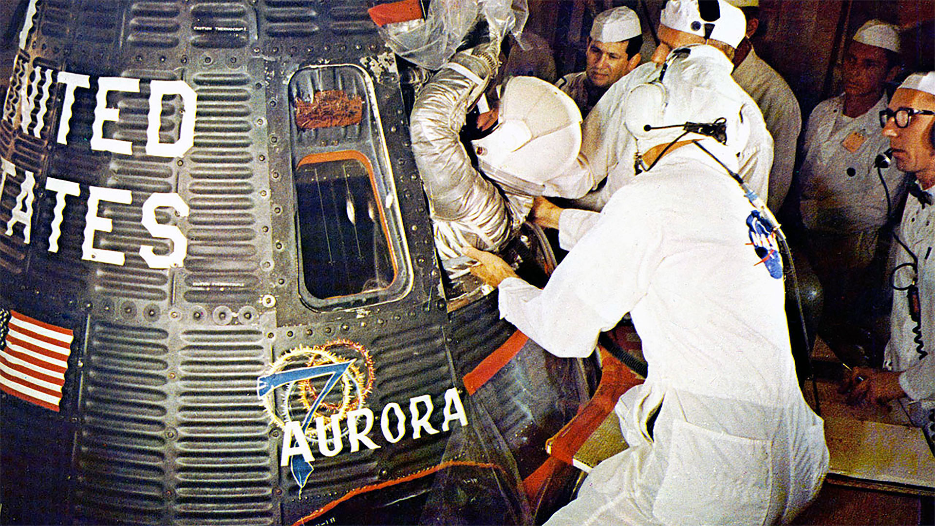 technicians in white clean room suits help an astronaut in a silver spacesuit and white helmet climb into a spacecraft for launch