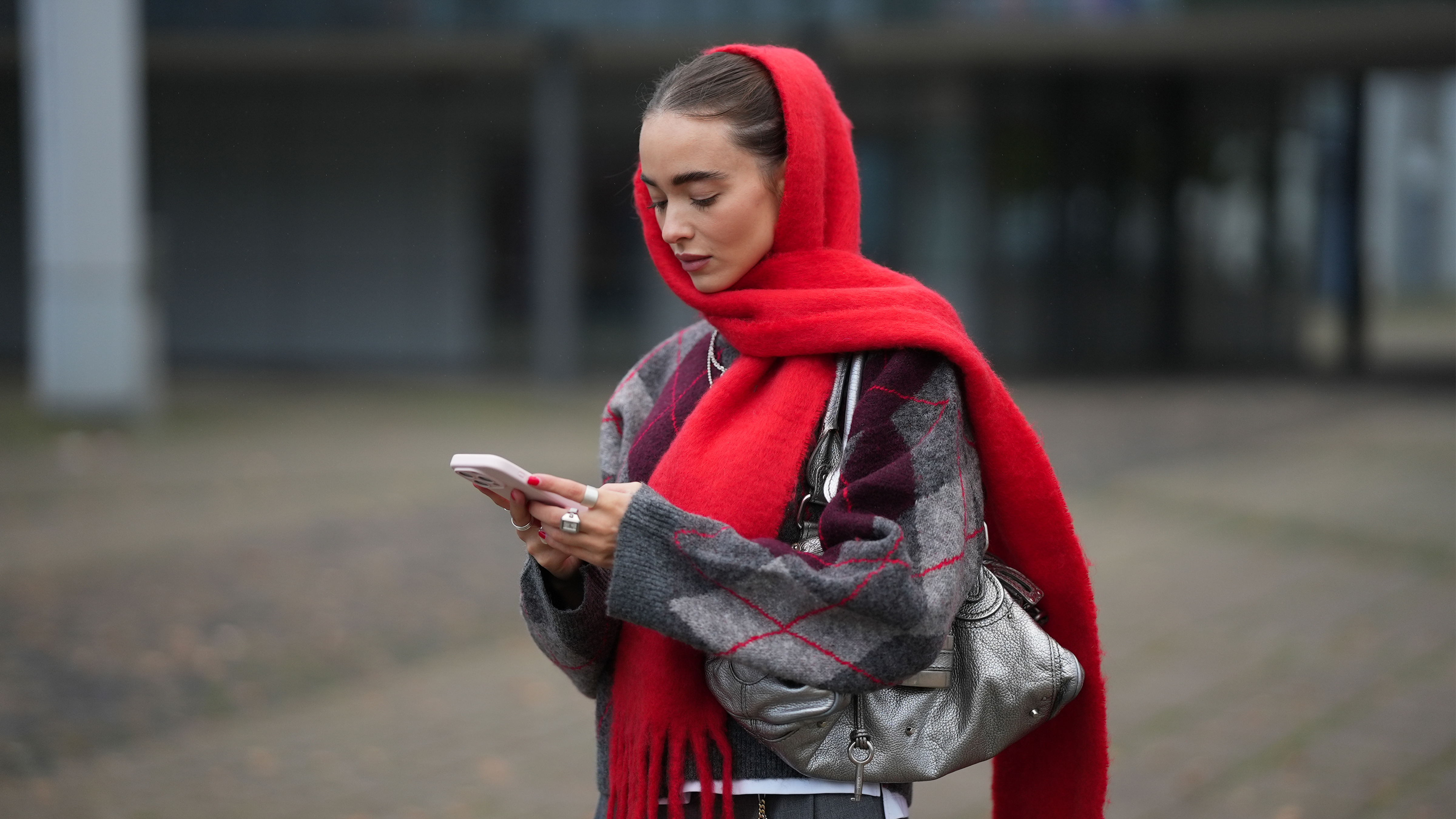 Woman in red knit scarf, argyle print sweater, silver metallic bag, and grey trousers, on her phone. 