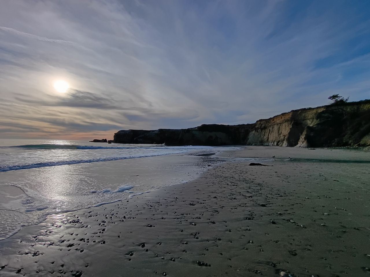ultrawide view of an empty beach next to a cliff with the tide out, water marks on the sand and the sun appearing behind slight cloud