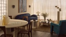 A living room with a blue sofa, a green armchair, a white dining table, wood chairs, a yellow vase, a glass egg crate, a chrome coffee and side table, a mushroom lamp, and a stacked silver floor lamp