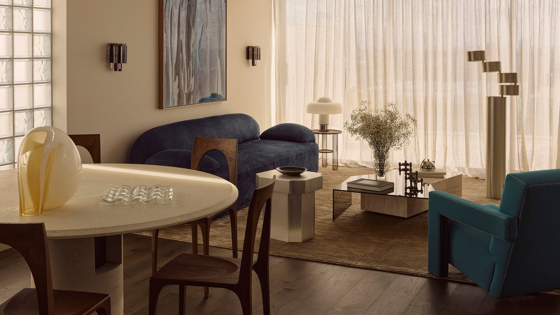 A living room with a blue sofa, a green armchair, a white dining table, wood chairs, a yellow vase, a glass egg crate, a chrome coffee and side table, a mushroom lamp, and a stacked silver floor lamp