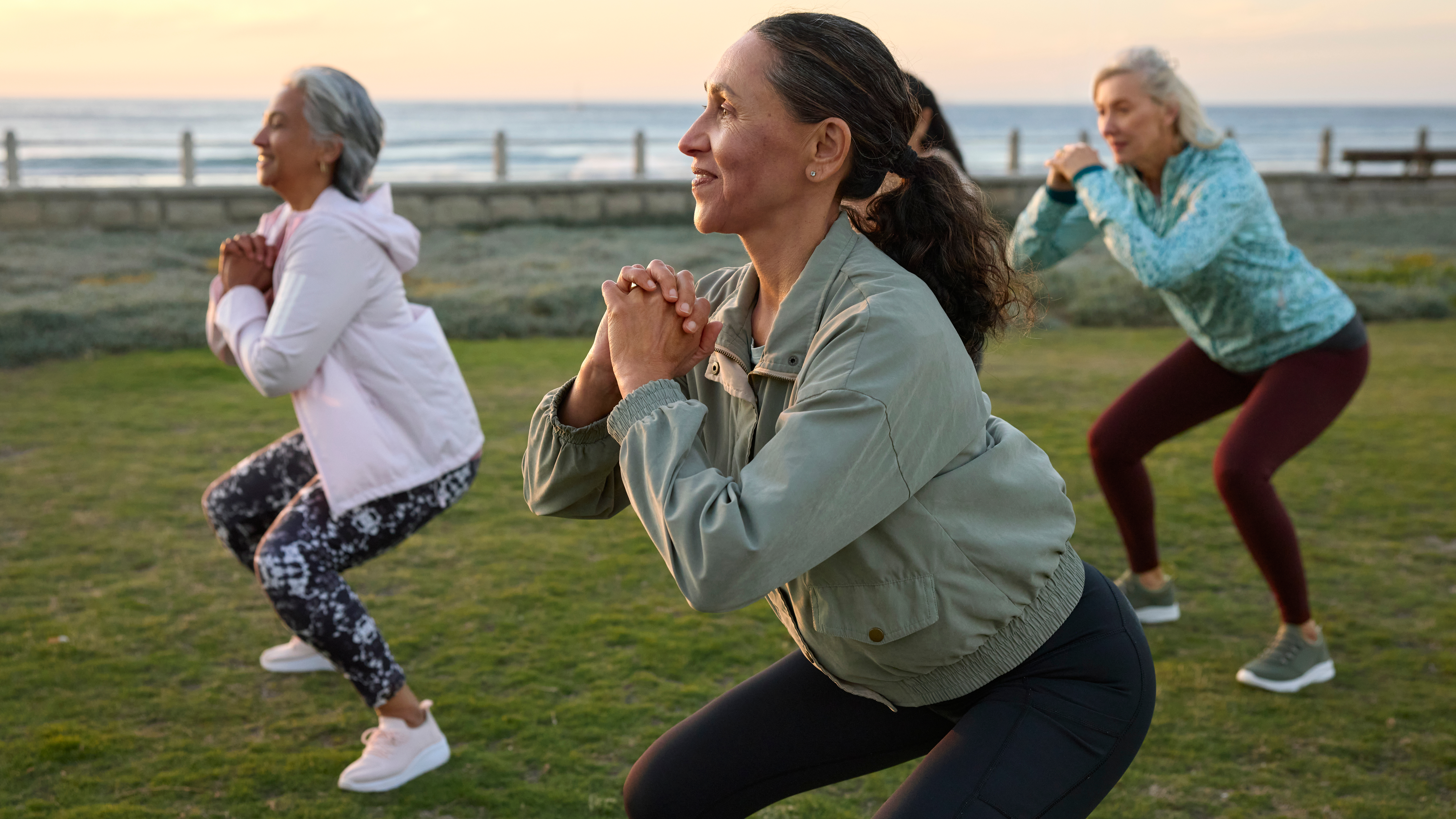 A group of women perform squats on a stretch of grass with the ocean behind them.