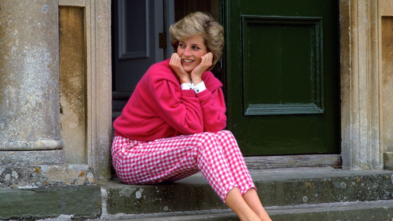 Princess Diana in a pink cardigan and gingham trousers sitting on the steps of Highgrove House