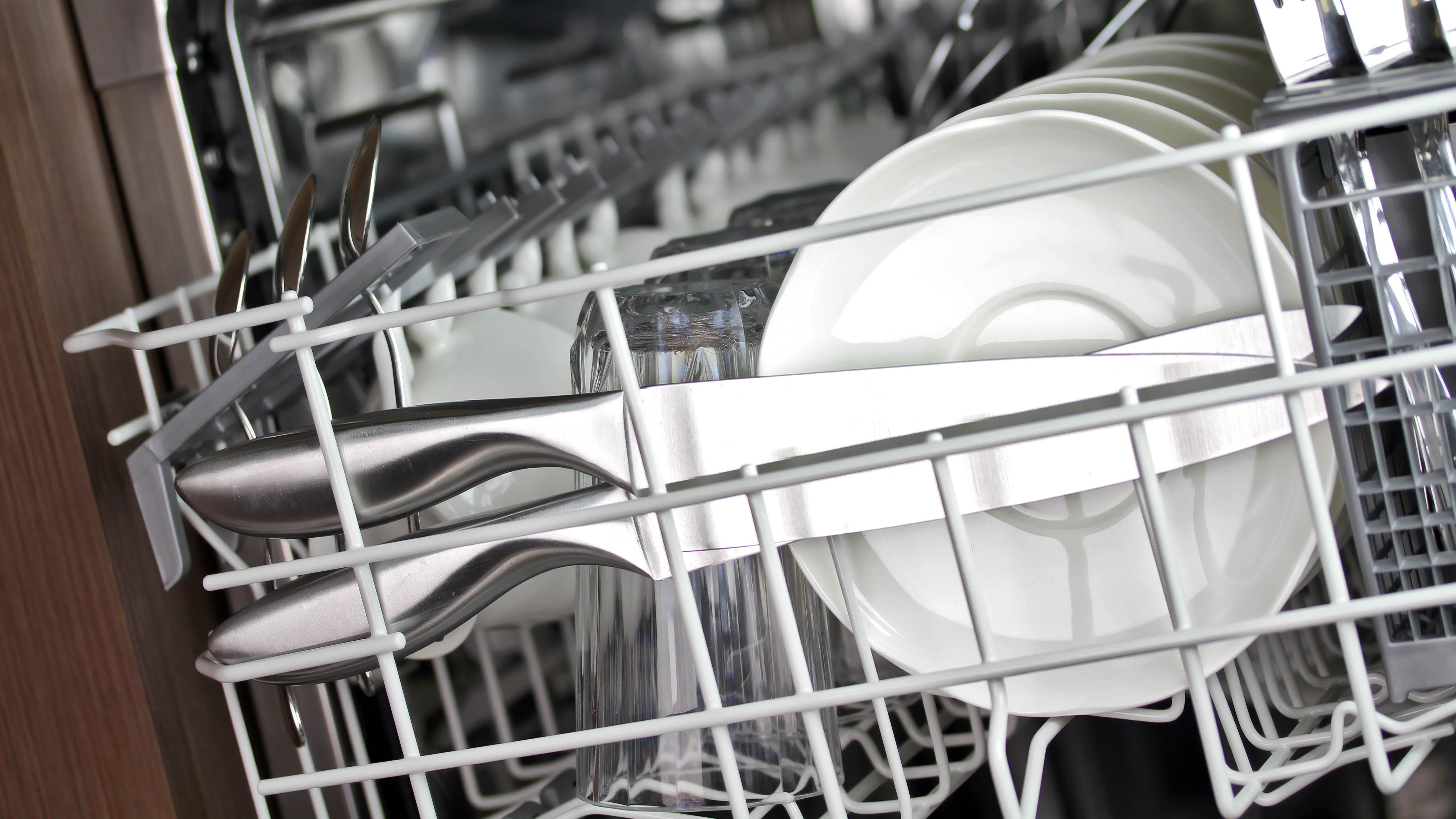 The upper rack of a dishwasher with kitchen knives in it next to plates
