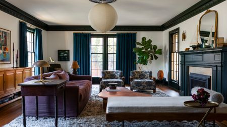 Living room with green ceiling trim, large windows with blue drapes, a large fireplace with a boat on the mantel, a purple sofa, a chaise lounge, and two floral armchairs, with a lantern light above.