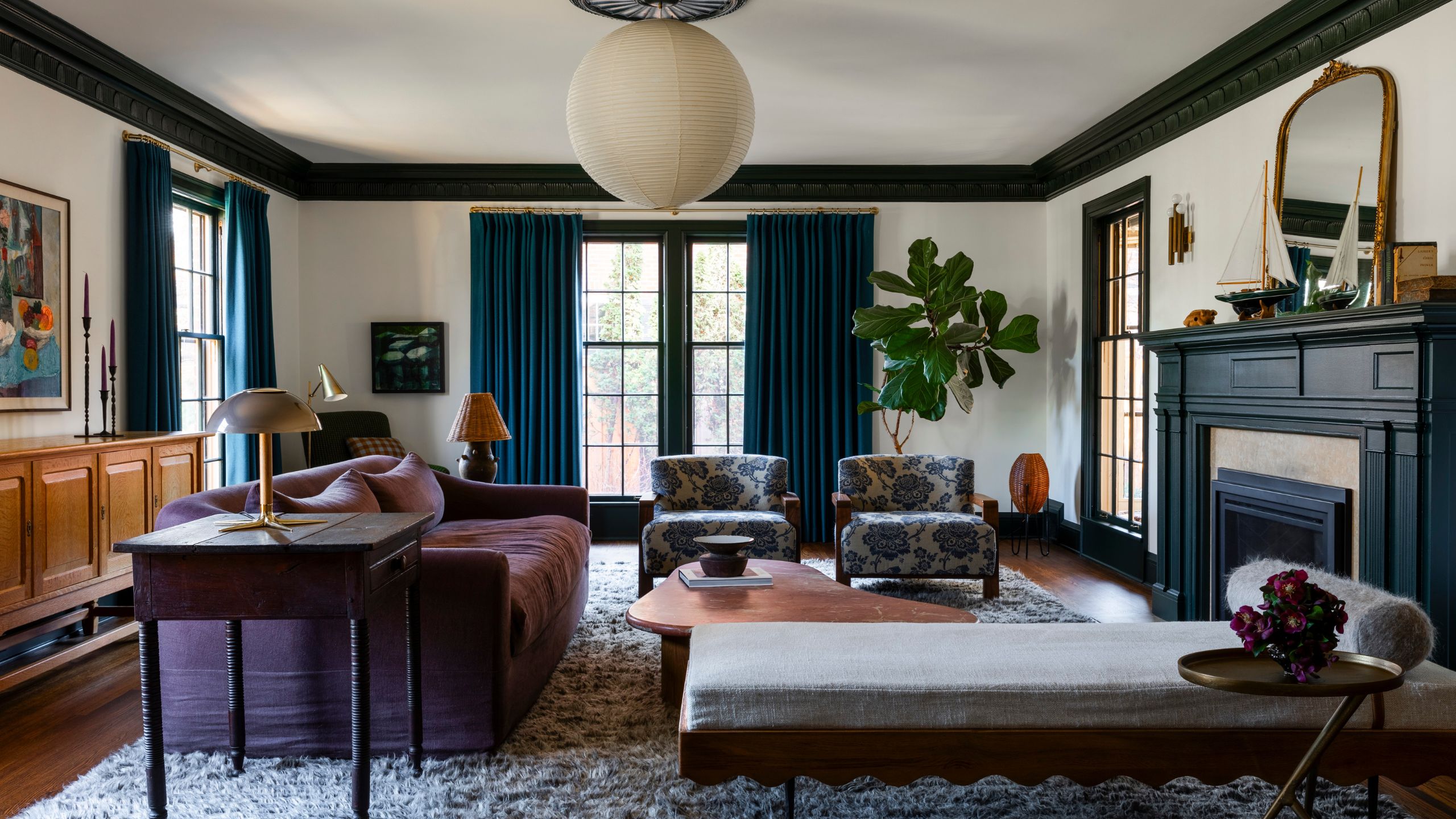 Living room with green ceiling trim, large windows with blue drapes, a large fireplace with a boat on the mantel, a purple sofa, a chaise lounge, and two floral armchairs, with a lantern light above.