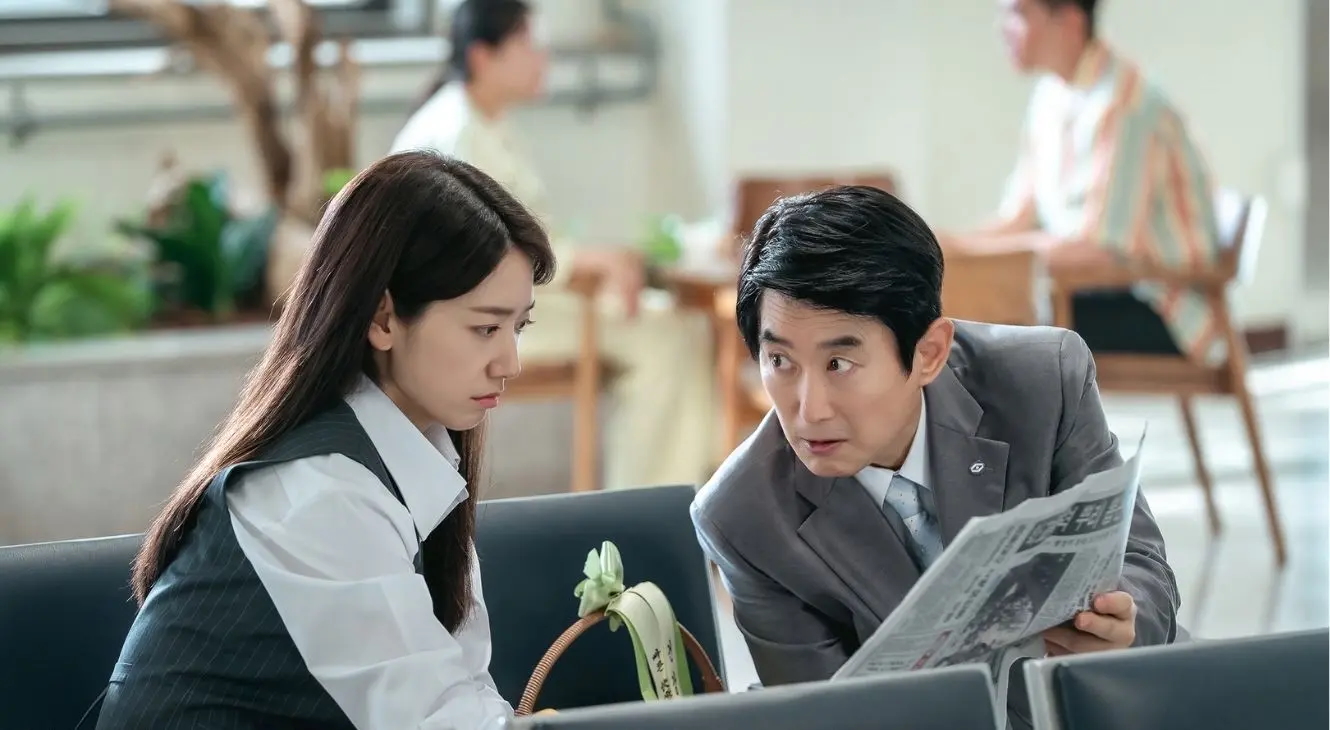 A woman (Park Shin-hye as Hong Keum-bo) leans toward a man (Kim Won-hae as Yun Jae-beom) as he holds up a newspaper. The pair sit in a hospital waiting area, in the Netflix K-drama 'Undercover Miss Hong.'