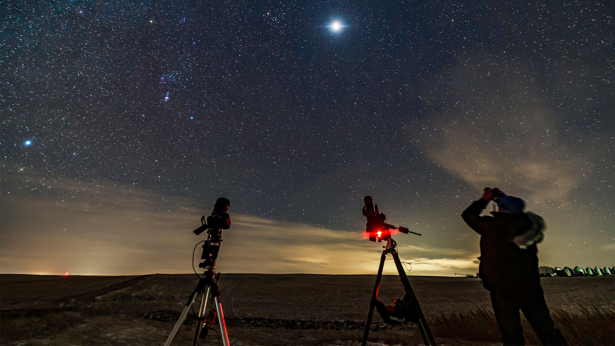 A person looking at the night sky through binoculars with telescopes also set up.