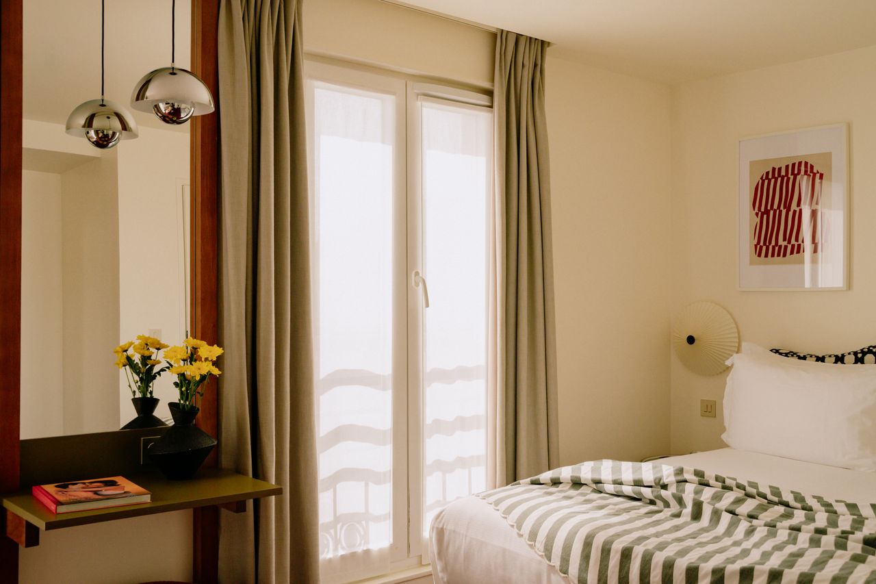 A sun-lit hotel room features an undone bed covered in a stripy throw, framed abstract wall art, a chrome pendant light, a book, and a vase of flowers.