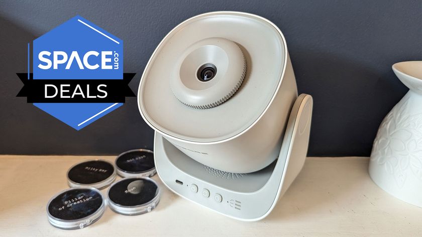 A white projector is balanced on a wide-based stand with buttons on the front, next to four black disks in a light wooden surface and in front of a grey background.