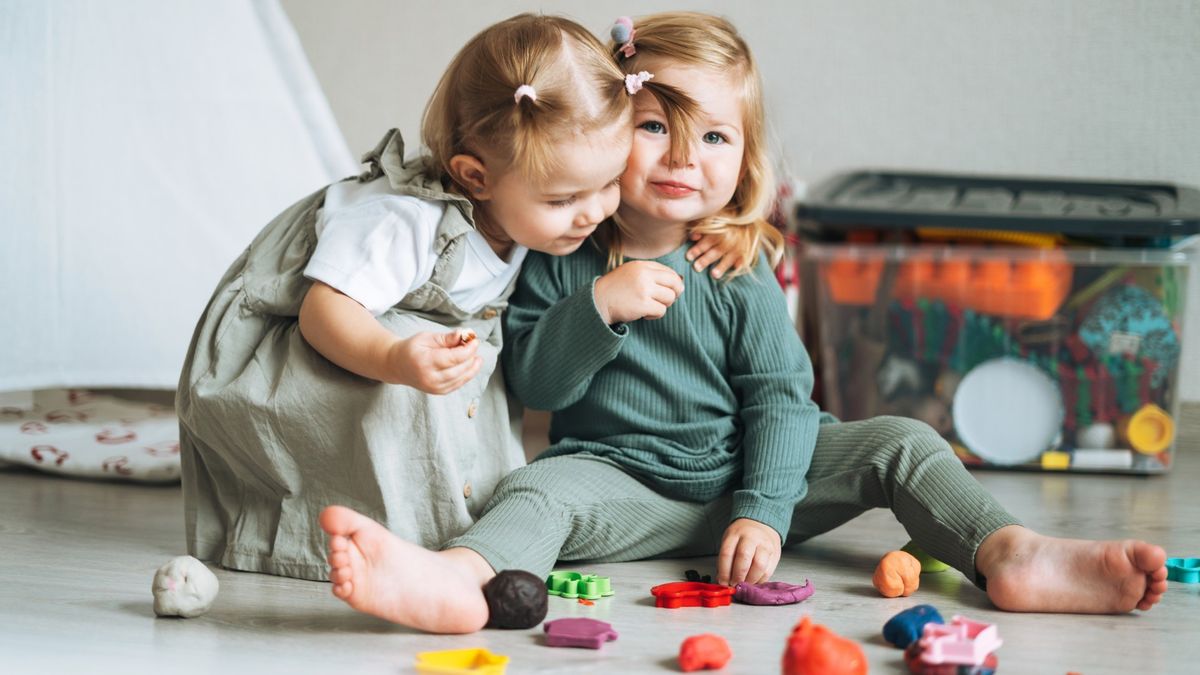 Two little girls are shown sat on a playroom floor with some toys in front of them. The girl on the left of the image has her arm around the other girl and is looking down at the floor. The girl on the right is looking at the camera.