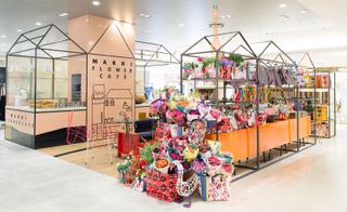 The Marni Flower Cafe. Flowers and gift bags displayed in metal frames on one side and food displayed in metal frames on the other side with tables, chairs and a sofa between them on a wooden floor.
