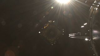 a gold cube-shaped satellite flies into the empty blackness of space beneath the sun, which causes bright rays across the camera lens viewing it