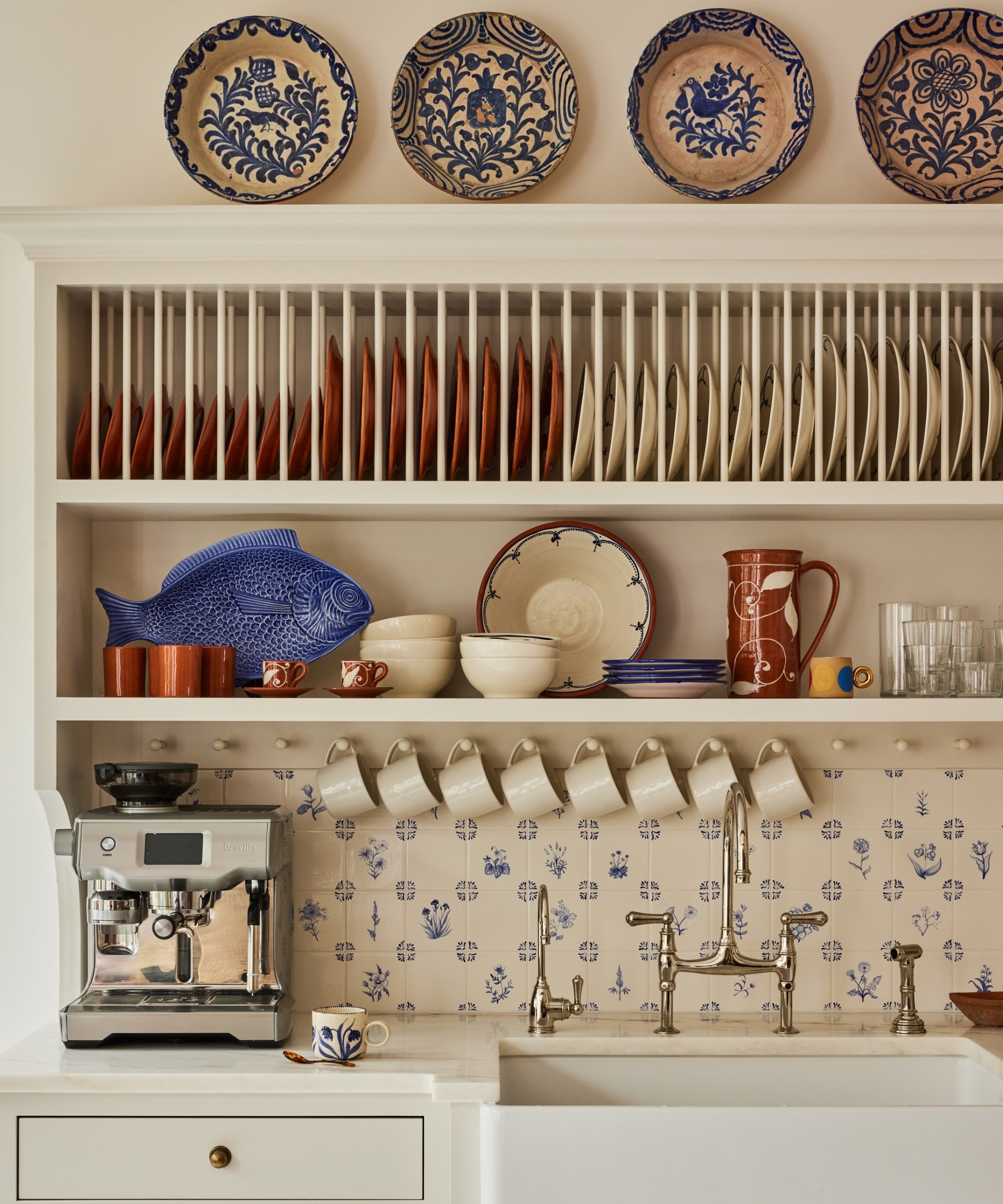 A close up of a custom hutch painted warm white with a plate rack at the top, open shelving below, and a kitchen sink, white countertops used as a coffee station and mugs hanging from a pegboard
