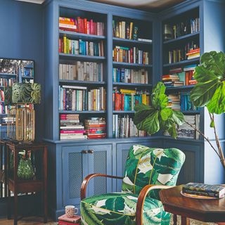 A built-in library painted the same shade of blue as the wall with a tropical-print accent chair in front of it