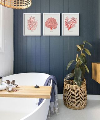 A bathroom with a blue full height slatted wooden wall, coral art prints, and a bath with a wooden tray