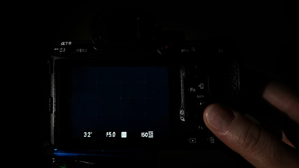 A close-up of a camera's rear screen displaying settings, with a hand adjusting controls in a dark environment
