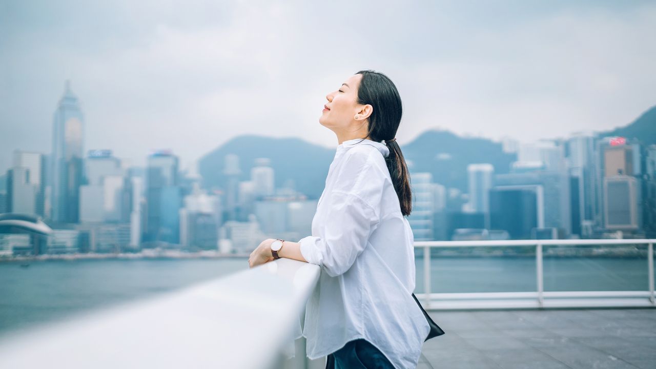 A young woman looks relaxed as she stands at a railing near a river with a city skyline in the background.