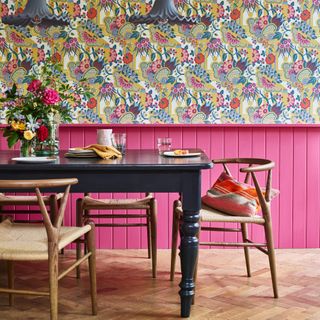 colour ideas for cottagecore decor, maximalist dining room with bright pink tongue and groove up to chair rail, stylised wallpaper above, flowers on dark grey dining table, blond wood chairs, grey pendant lights