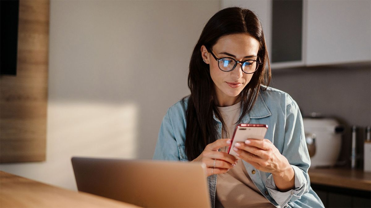 A professional woman browses her handheld device next to her laptop