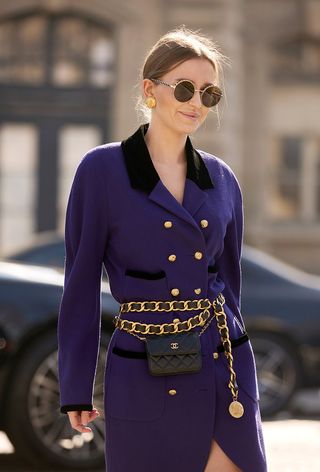 One of the best designer belts is shown in a close street-style photo of a woman wearing sunglasses, gold earrings, a purple blazer dress with a Chanel chain belt layered on top