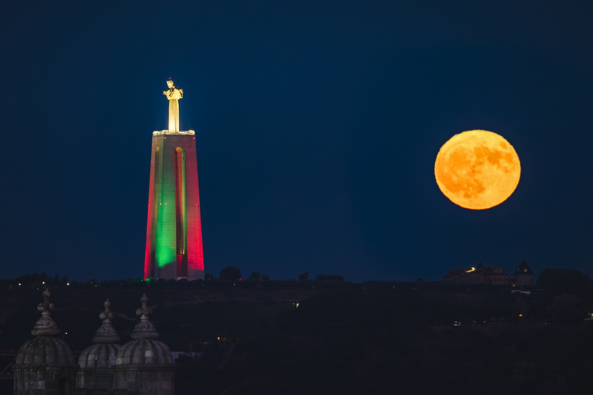 uma grande estátua de Cristo à esquerda é iluminada com luzes verdes e vermelhas enquanto a lua cheia brilha intensamente no céu à direita da estátua.