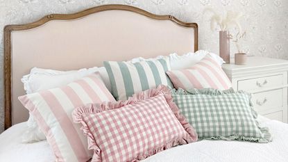 Headboard ideas are so dreamy. Here is a light pink headboard with a dark wooden trim with white sheets on it and pink and green gingham and striped throw pillows on it