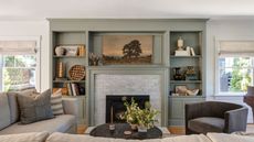 Living room with grey-green painted built-in bookshelves with decor and books, fireplace with a landscape painting above it, neutral sofa, and a dark grey velvet armchair 