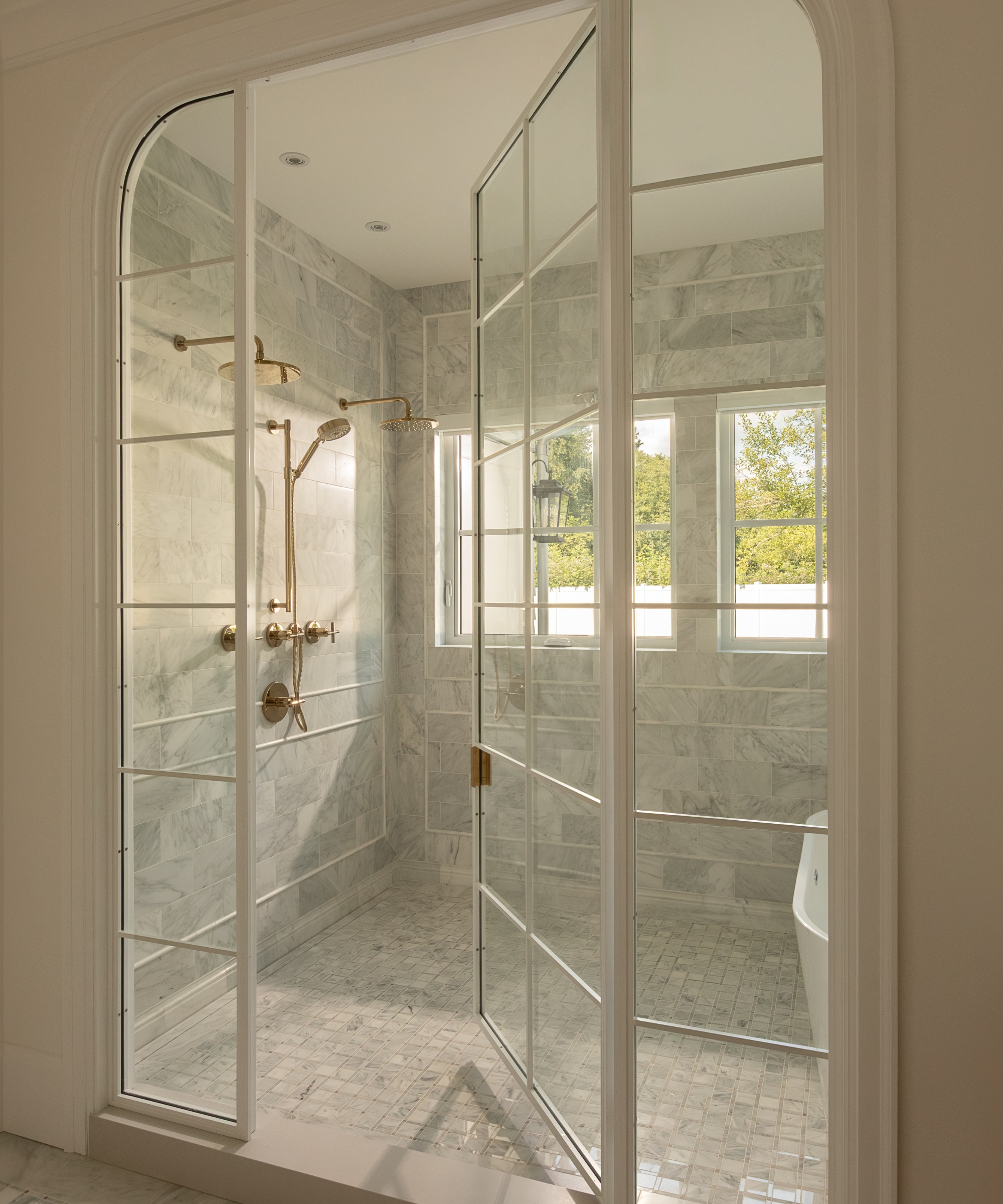 A wet room with glazed arched doors, marble tiles, brass hardware, and a freestanding bath