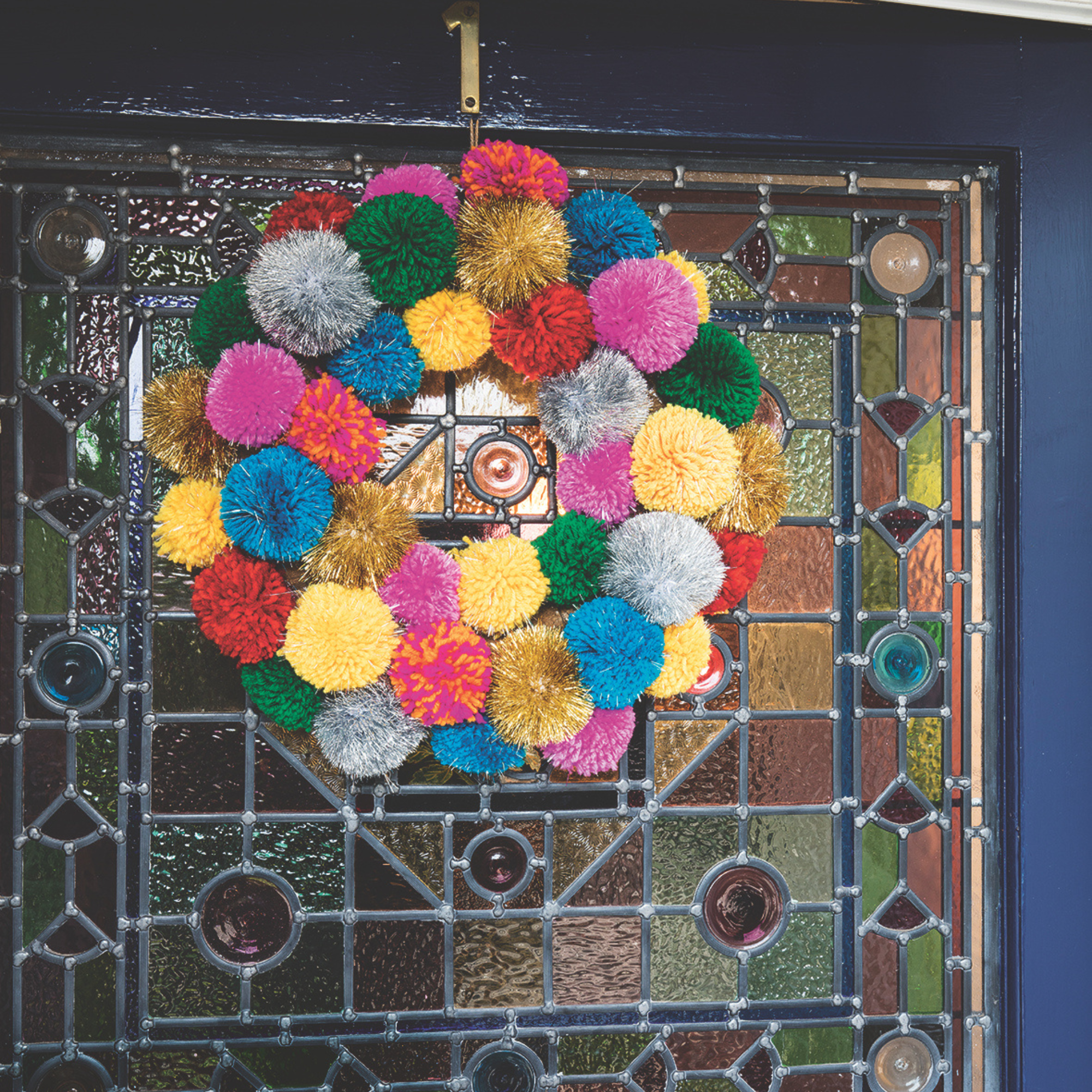 Wreath of colourful pompoms hanging on the front door in front of a stained glass panel.