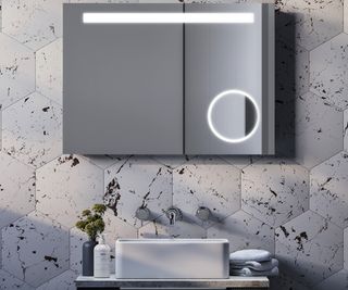 mirrored bathroom cabinet with LED light across top of doors and lit magnified section on bottom right hand of one door on tiled wall above white sink and wall mounted taps