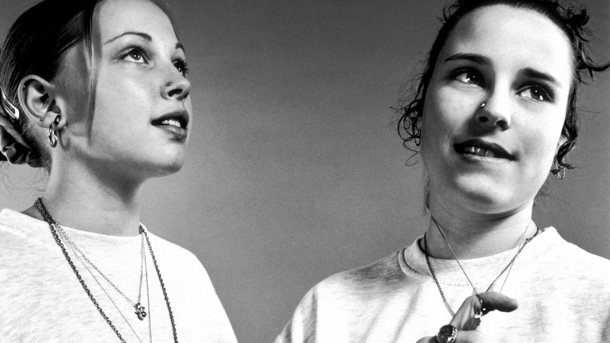 Black-and-white photograph of two young women wearing white Reebok T-shirts, layered necklaces and hoop earrings, looking off-camera.