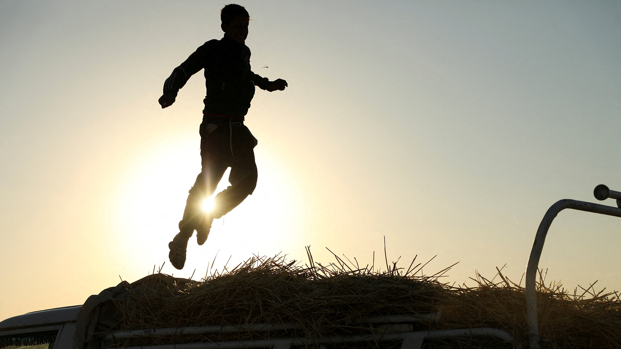 A boy is silhouetted as he jumps on top of a truck loaded with straw after the wheat harvest in Qamishli, Syria