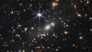 A view of space with lots of specks of light indicating galaxies and a few streaks representing lensed light.