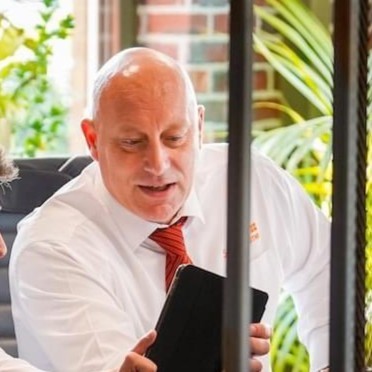 Bald man in white shirt, red tie, speaking while holding a tablet at an outdoor café table with plants behind him.