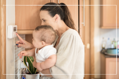 Young Mother holds her baby boy on her arms and shows him how to adjusts the temperature of the household on a thermostat in the kitchen