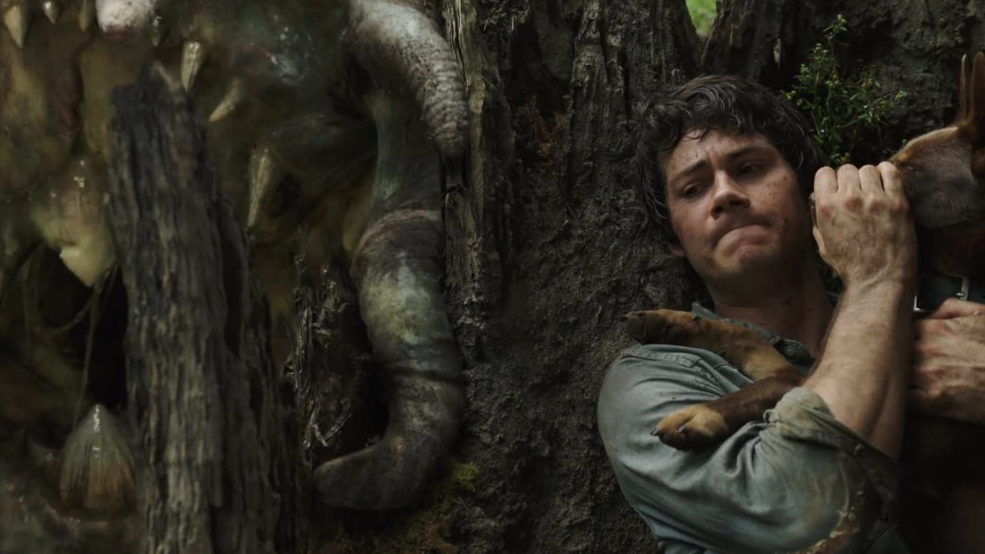 Dylan O'Brien as Joel hiding from a large creature behind a tree in the Netflix movie, Love and Monsters.