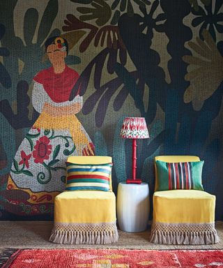 Hallway with bold hanging mural with frida kahlo in woodland scene, two bright yellow upholstered chairs with colorful cushions, side table with red lampshade in-between, red patterned rug