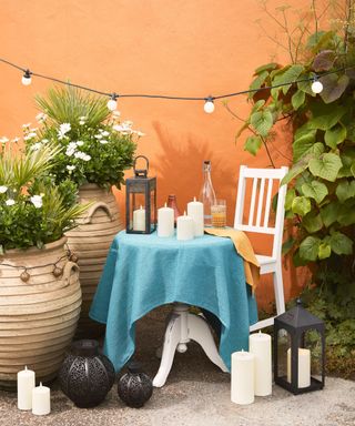 An outdoor seating area with a bright orange wall, a table with a blue cloth with candles on top and a white chair next to it, lanterns and candles below it, and pots of plants around it