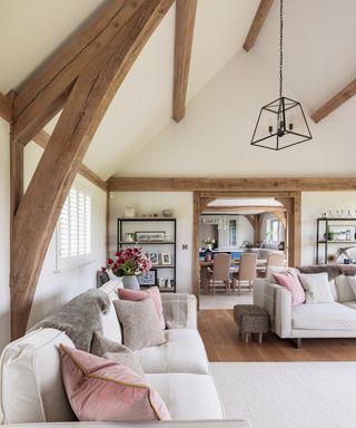 A cosy living room in an oak frame self-build home. The beams are partially exposed giving a clean yet rustic finish. The interiors are neutral with two sumptuous cream sofas with plump blush and oatmeal cushions and a cream carpet sits on a laminate wood flooring. There is a framed entranceway in the background through which a dining room table can be glimpsed