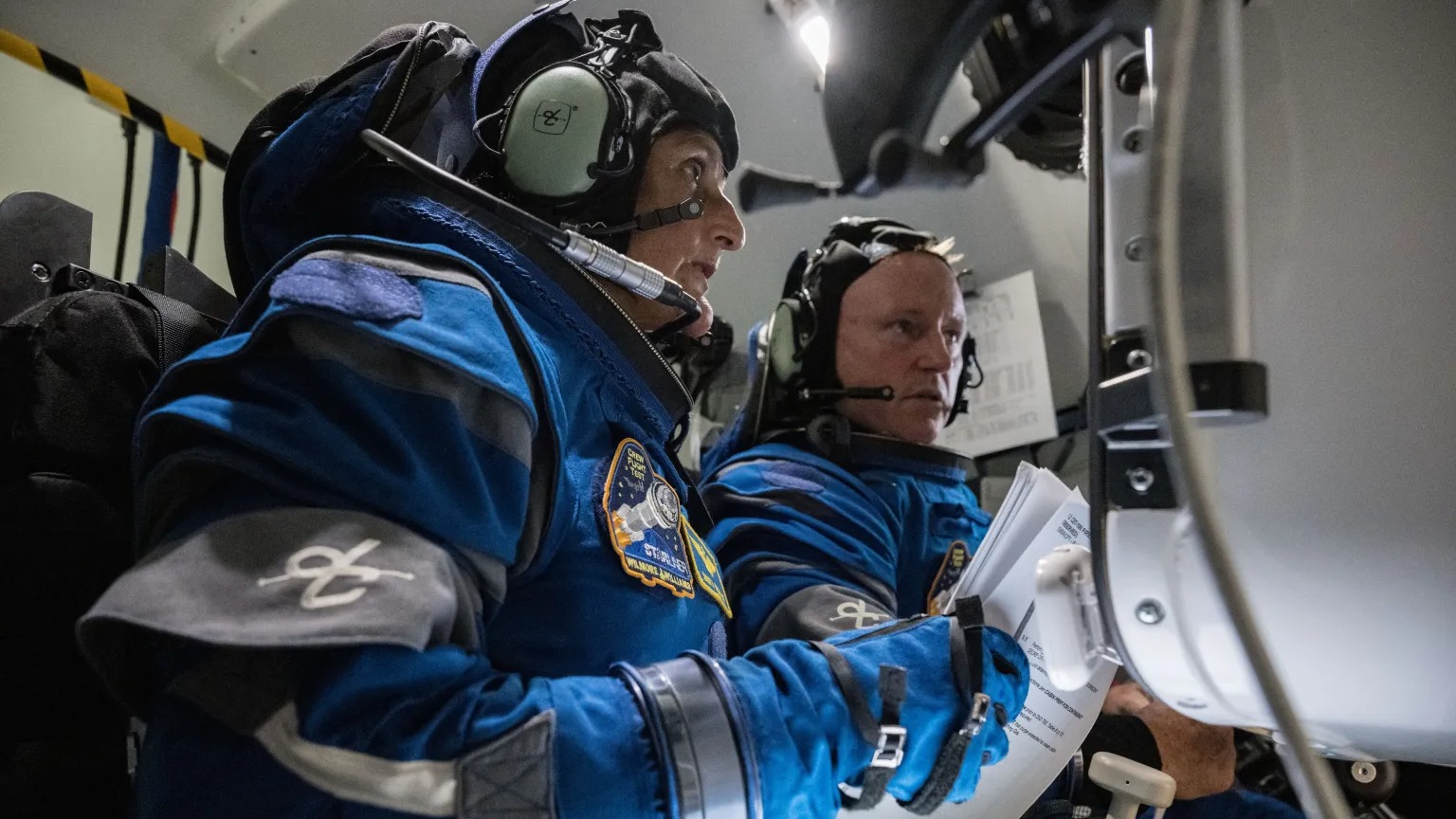 two astronauts in training in a simulator with spacesuits on, looking at a screen