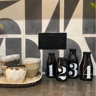 black double plug socket against grey and white tiles with black bottles and cream and beige bowls