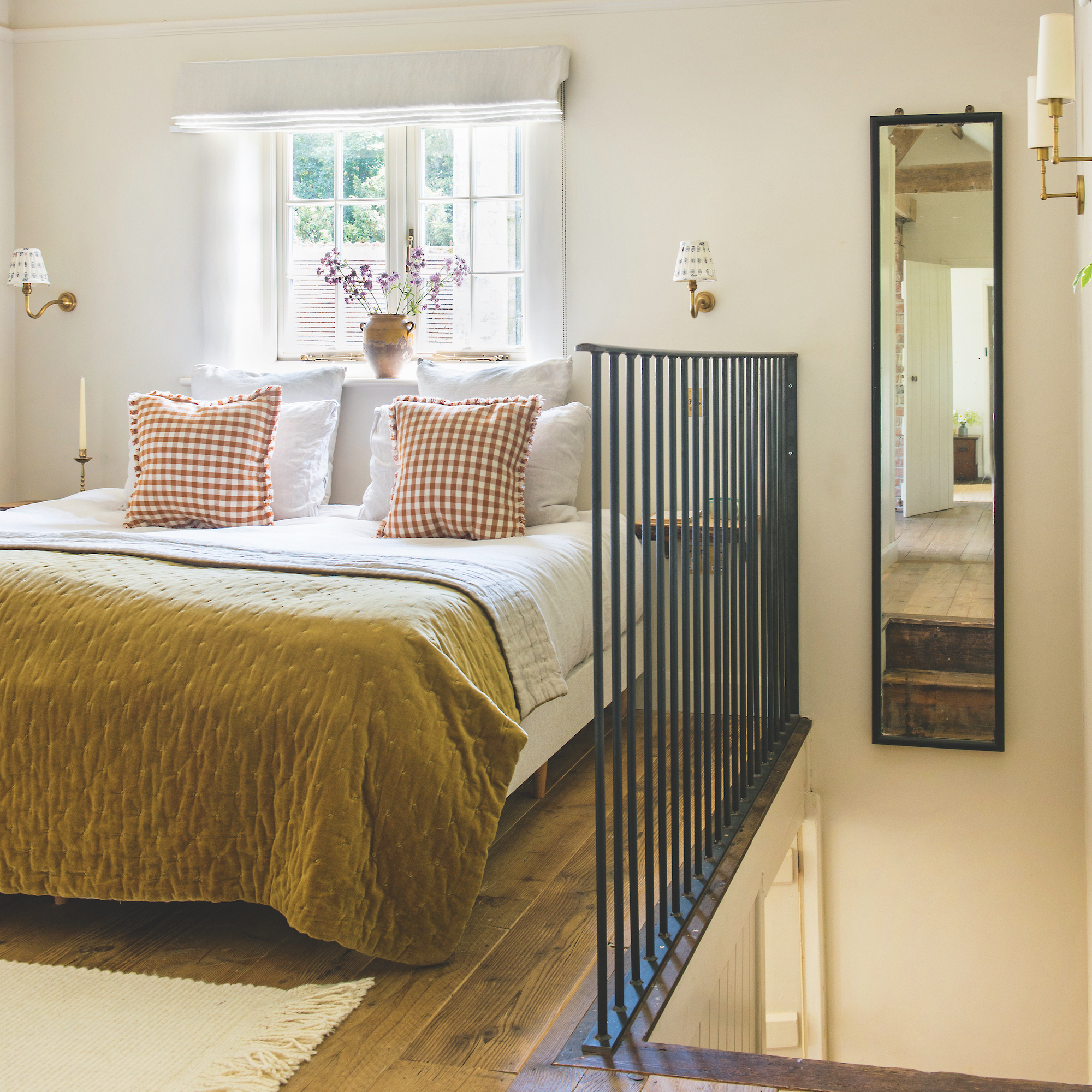 bedroom with white walls and metal rail next to staircase, check cushions and gold velvet bedspread