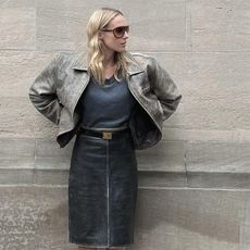 Elly wears a black top and black leather skirt with a brown. jacket while turning head and wearing sun glasses