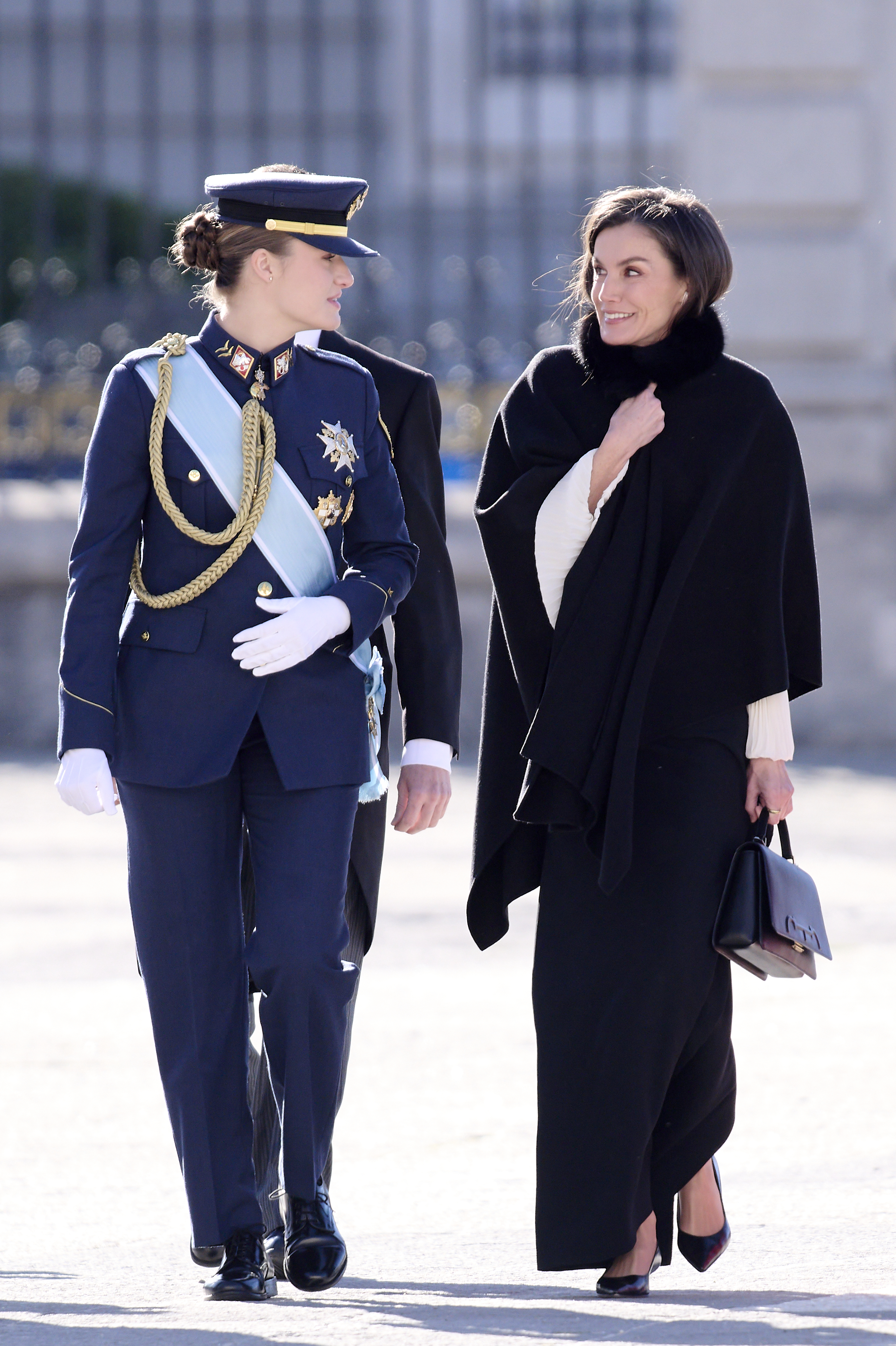 Princess Leonor wearing a blue military uniform walking next to Queen Letizia clutching a black cape