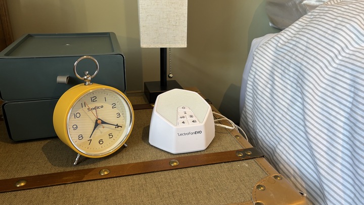 The LectroFan EVO White Noise Machine next to a white lamp and a yellow alarm clock on a nightstand.