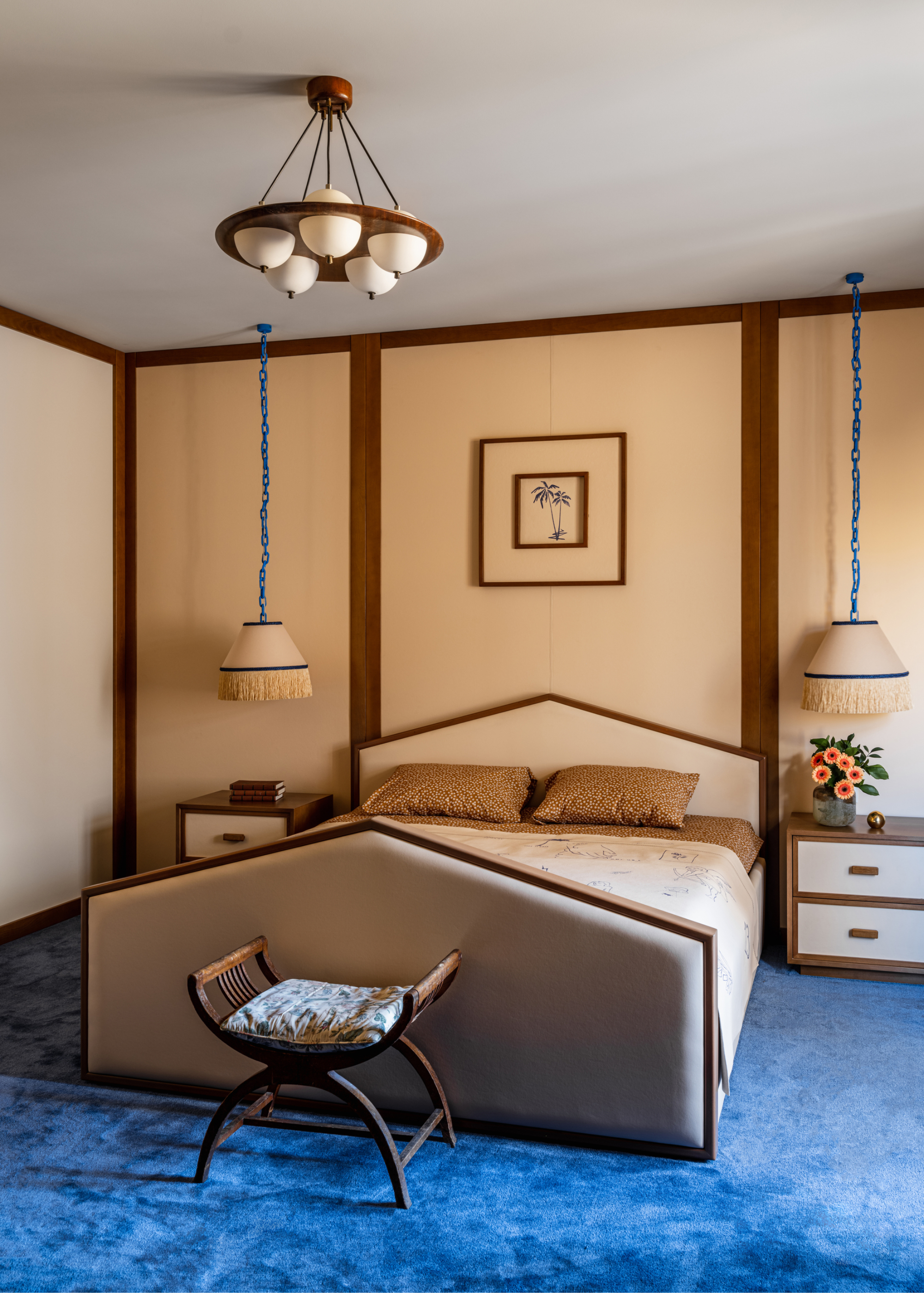 bedroom with bright blue carpet, beige wall panels, upholstered bed frame, seat at end of bed, two side tables with pendant lights hanging above