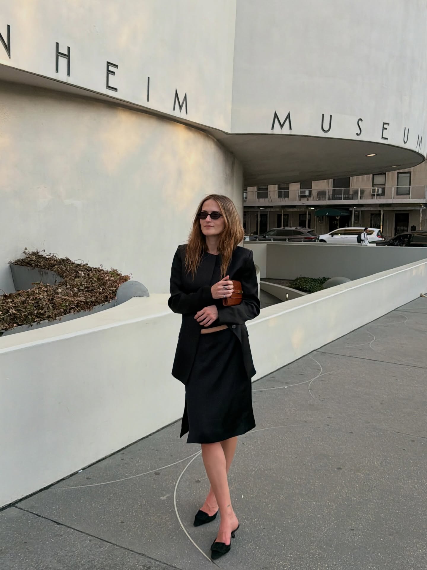Eliza Huber wearing a satin skirt suit with a brown clutch and black mules.