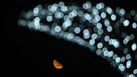 A half-lit moon shines beneath out-of-focus artificial lights arrayed in an arc against a black sky.