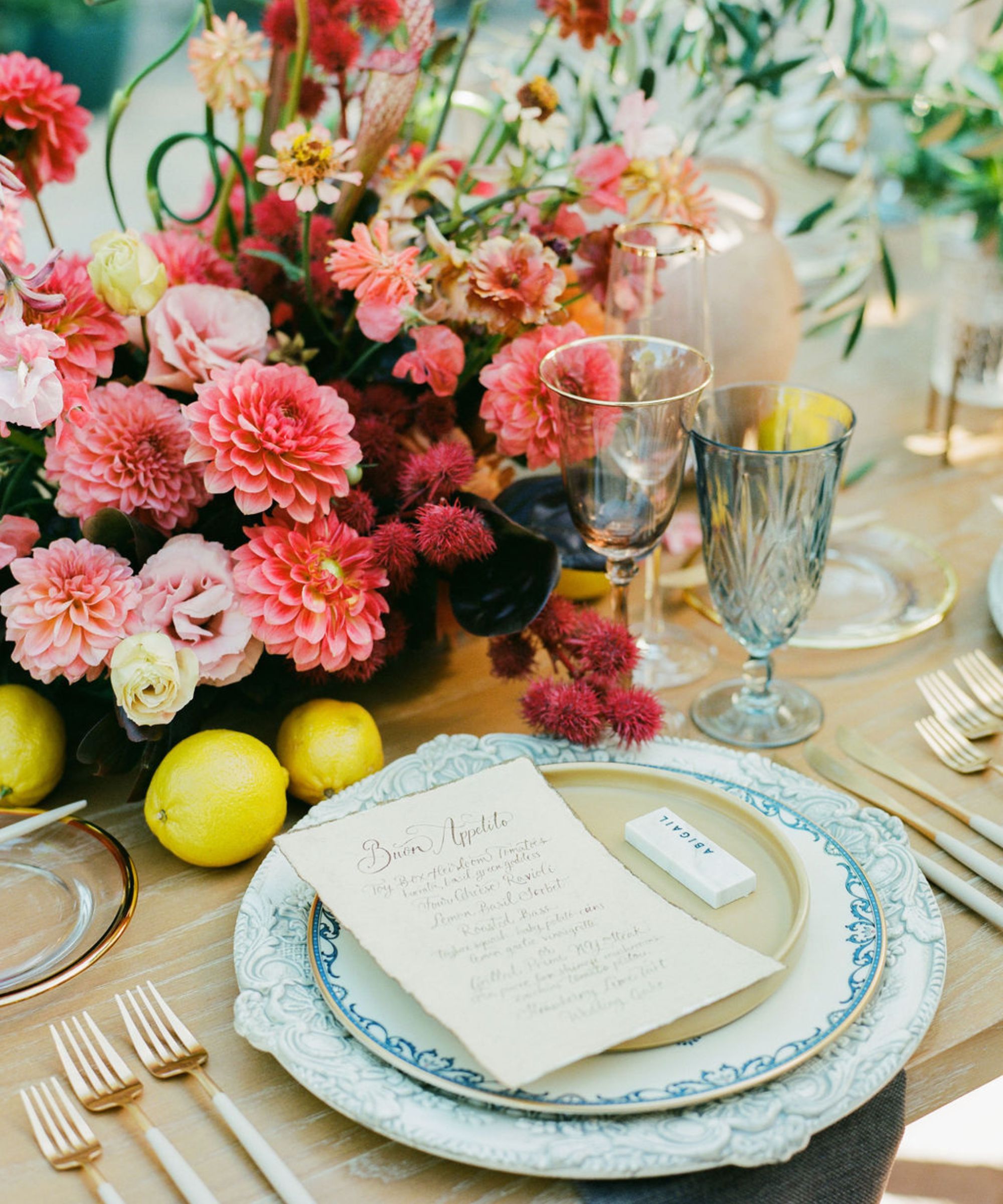 A tablescape with pink dahlias and flowers at the centre, blue china plates and gold cutlery, lemons scattered on the table, and blue glassware